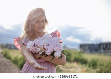 Charming woman with disheveled hair in summer sundress with luxurious bouquet of peonies on summer day stretches her hand forward. Femininity and beauty. Positive emotions. Growing flowers. - Powered by Shutterstock
