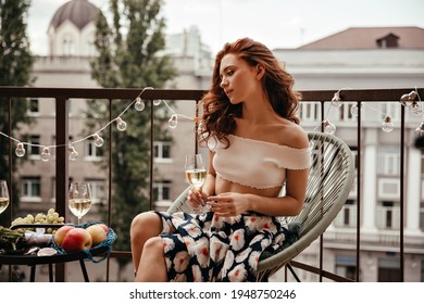 Charming woman in floral outfit holds wine glass and rests on terrace. Red-haired girl with champagne sits on white modern chair and relaxes. - Powered by Shutterstock