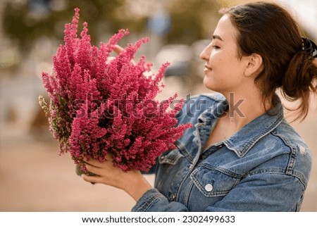 charming woman holds large flower pot with growing flowering pink heather plant in the hands.