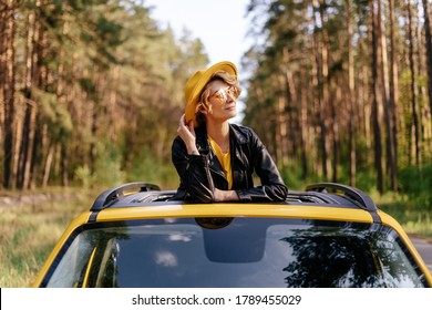 Charming Woman Leaning At Yellow Auto Sunroof. Dreamy Girl In Leather Jacket, Hat And Sunglasses Standing Out Of Car Roof Front View. Caucasian Stylish Model Daydreaming Looking At Forest