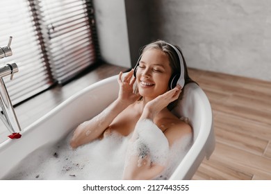 Charming woman lying in hot bubbly bath and listening to music in wireless headphones, free space. Pretty young lady enjoying favorite playlist on domestic spa day