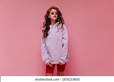 Charming Woman In Red Cycling Shorts And Purple Hoodie Poses On Pink Background. Young Girl In Sunglasses Looks Into Camera On Isolated.