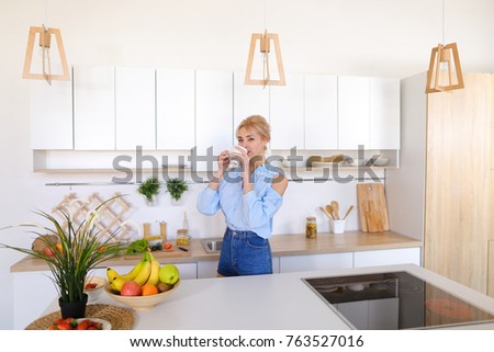 Charming woman walks up and goes to kitchen table, raises hands to sides and up to relieve tension and takes in hands fragrant morning fresh coffee, smiles with easy smile, standing in middle of