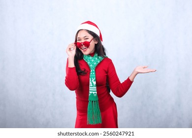 charming woman wearing red sweater with Santa hat and scarf, cheerful to camera hands holding red glasses and presenting to left, celebrating merry christmas and new year isolated on white background - Powered by Shutterstock