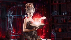 Charming Woman Witch Flowers In Hair Vintage Black Dress With Book Of Spell In The Old Castle. Halloween. Celebration. Witchcraft Concept, Fairy Tale Mystic Atmosphere. Conjure Magic Spells