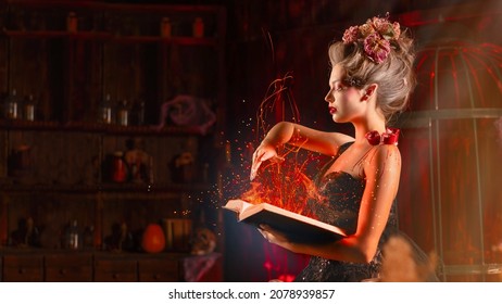 Charming Woman Witch Flowers In Hair Vintage Black Dress With Book Of Spell In The Old Castle. Halloween. Celebration. Witchcraft Concept, Fairy Tale Mystic Atmosphere. Conjure Magic Spells