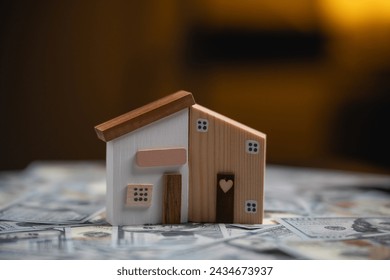 Charming Wooden House Model on Cash Background. A quaint wooden house model stands on a background of scattered US dollar bills. Finance home loan or interest concept. - Powered by Shutterstock