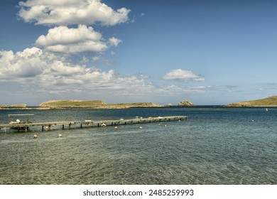 A charming wooden pier stretching into the clear waters of Puerto Addaya, Menorca, with a scenic view of green hills under a bright blue sky with wispy clouds. - Powered by Shutterstock