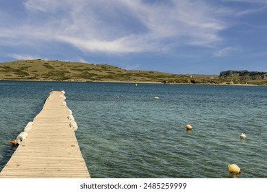 A charming wooden pier stretching into the clear waters of Puerto Addaya, Menorca, with a scenic view of green hills under a bright blue sky with wispy clouds. - Powered by Shutterstock