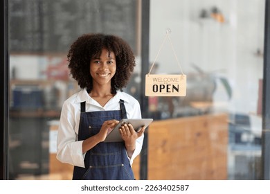 Charming young African American businesswoman owner wearing an apron holding a tablet in front of a coffee shop counter.  - Powered by Shutterstock