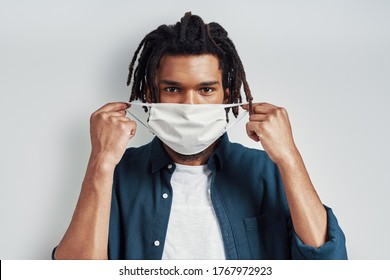 Charming young African man adjusting medical face mask and looking at camera while standing against grey background - Powered by Shutterstock