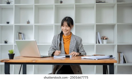 A charming, young Asian businesswoman in a trendy suit is working at her desk in the modern office, reviewing business reports and looking at her laptop screen. - Powered by Shutterstock