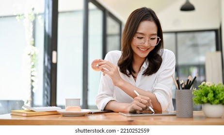 Charming young Asian female office employee or college student using a digital tablet to design her artwork while enjoying her yummy doughnuts at the cafe. - Powered by Shutterstock