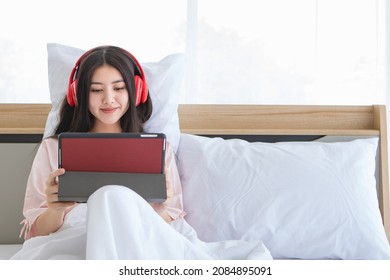 Charming Young Asian Girl Happy To Rest On Bed And Enjoy Online Music Listening And Entertainment Media From Modern Tablet By Comfortable Earphone Gadget While Easily Leaning On Pillow On Holiday.