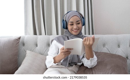 Charming young Asian Muslim woman wearing hijab and headphones, listening to music and reading a book in the living room.
 - Powered by Shutterstock