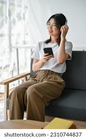 A charming young Asian woman in casual clothes enjoys the music on her headphones while sitting on a sofa in her living room. - Shutterstock ID 2310744837