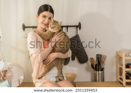 A charming young Asian woman sits on a kitchen counter, holding her adorable cat tightly as morning sun streams in