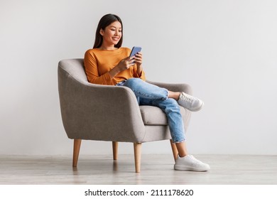 Charming young Asian woman using cellphone, communicating on web, working or learning online, sitting in armchair against white wall, free space. Lovely millennial lady chatting on smartphone