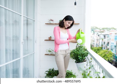 Charming Young Asian Woman Watering Plant In Container On Balcony Garden