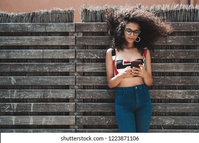 Charming young biracial female leaning against a wooden hedge with wattle fence inside and checking incoming messages on her smartphone, with a copy space area on the left for your text or logo - Powered by Shutterstock