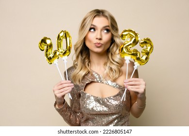 Charming young blonde girl with curly hair wearing sparkle dress holding balloons number 2023 in her hands on a beige background, new year holiday concept