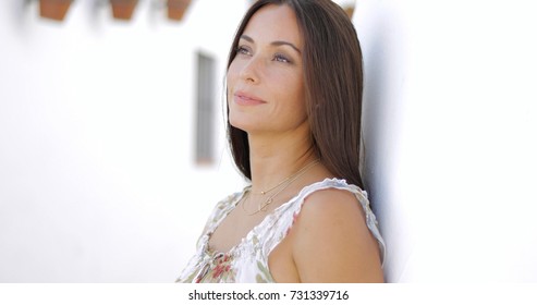 Charming young brunette in light dress leaning on street wall while looking away and daydreaming happily.