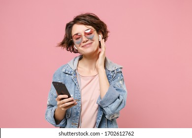 Charming Young Brunette Woman In Casual Denim Jacket, Eyeglasses Posing Isolated On Pastel Pink Background. People Lifestyle Concept. Mock Up Copy Space. Listen Music With Air Pods, Hold Mobile Phone