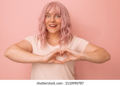 Charming young caucasian pink hair woman feels happy and romantic shapes heart gesture poses against pink background.