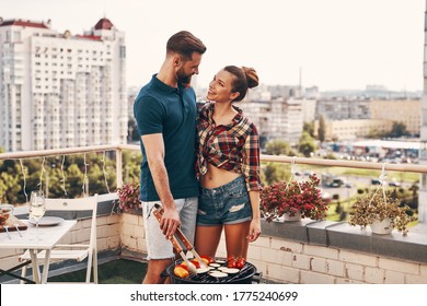 Charming young couple in casual clothing embracing and smiling while standing on the rooftop patio outdoors   