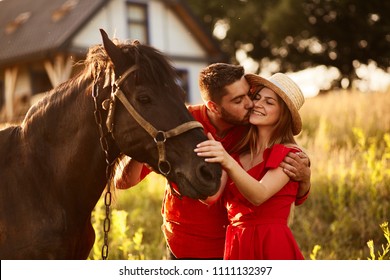Charming young couple stands with a brown horse before a country house in the rays of evening sun