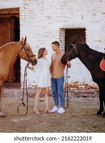 Charming young couple stands with two horses before a country house on the ranch