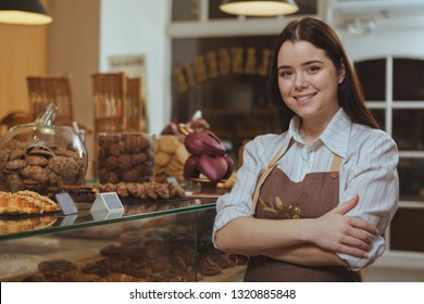 Charming young female baker smiling to the camera, posing proudly at her own bakery cafe. Happy female confectioner enjoying working at her bakery, copy space. Entrepreneurship concept