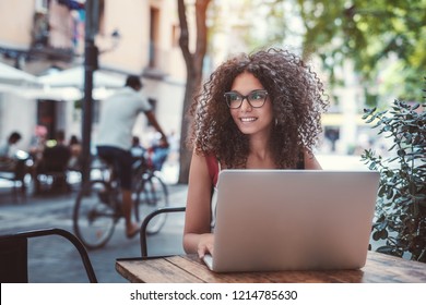 Charming young female caucasian freelancer in glasses is sitting in a street cafe with her netbook and looking aside; beautiful woman in eyeglasses in a bar outdoors with the laptop, a cyclist behind