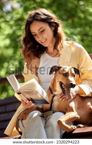 A charming young girl is resting on a bench in the park with a golden dog on sunny day. The girl reads a book and hugs her pet. Love and affection between owner and pet. Adopting a pet from a shelter.