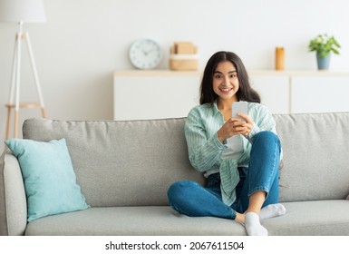 Charming Young Indian Woman Sitting On Sofa With Smartphone, Checking Social Media, Chatting Online At Home, Copy Space. Lovely Eastern Female Posting On Web, Watching Video On Mobile Device