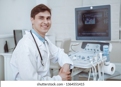 Charming young male doctor smiling to the camera, sitting at his office near ultrasound scanning machine. Handsome friendly doctor enjoying working at the hospital, copy space. Ultrasonic concept