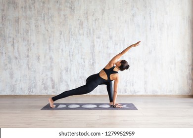 Charming Young Slim Woman Yoga Instructor Doing Parivritta Parsvakonasana Standing On A Rug On A Wooden Floor In A Spacious Hall With Daylight