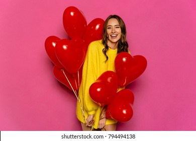 Charming young woman celebrating St. Valentine's day, holding red air balloons. Dressed in yellow sweater. Isolated over pink background.