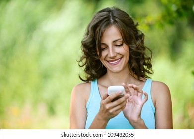 Charming Young Woman Close-up In Blue Shirt Reads Message To Mobile Phone, Against Green Of Summer Park.