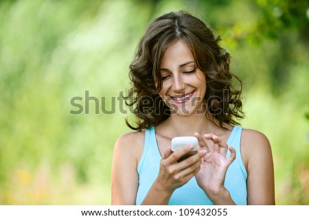 Charming young woman close-up in blue shirt reads message to mobile phone, against green of summer park.
