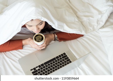 Charming Young Woman Covering Head With White Blanket Lying On Bed And Watching Tv Or Movies Streaming On Laptop And Holding A Cub Of Hot Coffee In Winter Season.