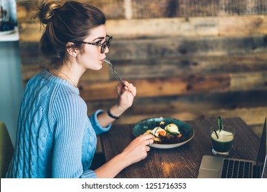 Charming young woman freelancer in stylish eyeglasses eating sitting at wooden table with plate of delicious food and drink in coffee shop.Hipster girl in eyewear have dinner in modern cafe interior