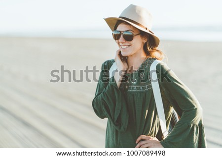 Charming young woman with hat, sunglasses, backpack with cute smile standing on the beach at sunset in warm weather. Boohoo style.
