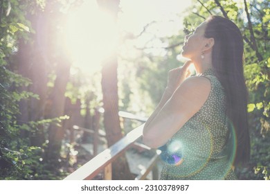 Charming young woman with long hair stands on path in forest and looks thoughtfully at sky. Dreamy lovely girl among nature. Beautiful sun rainbow highlights and reflections. - Shutterstock ID 2310378987