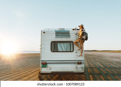 Charming young woman with nice smile with hat, sunglasses, backpack climing on recreational vehicle on the ocean beach at sunset. Stock Photo