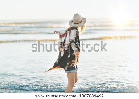 Charming young woman with poncho, hat with beautiful smile standing on the beach at sunset in warm weather.