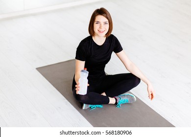Charming young woman relaxing after workout while sitting on the mat  at fitness gym. Pretty athletic girl with short brown hair holding bottle with water and smiling at camera. 