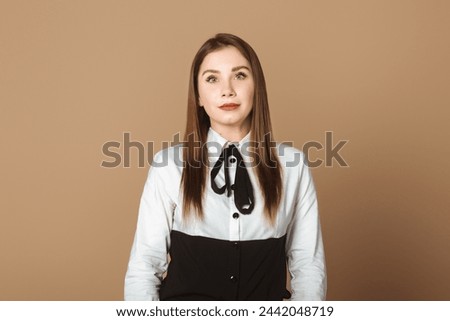 Charming Young Woman Standing Against Beige Studio Background, Gazing Assertively at the Camera. Stylish and Self-assured Female Portraiture in Neutral Setting. Beauty, Confidence and Elegance Concept