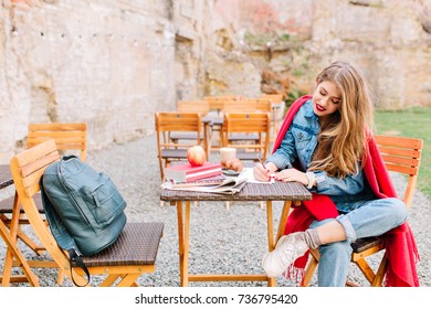 Charming Young Woman Wearing Stylish Denim Jacket, Enjoys Sunday Morning And Writes A Personal Letter Sitting In The Yard On Stone Wall Background. Positive Emotions, Writing Poetry, Sunny Day