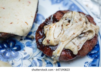 Charred Organic Grass Fed Beef Hamburger Patty Covered With Stringy Swirled Melted White Pepper Jack Cheese Dripping With Fat On Ornately Painted Blue And White Platter Plate With Cheese Burger